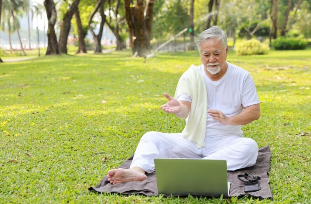A senior with mobility problems watches an online tai chi class and follows the movements while seated on a blanket outside.