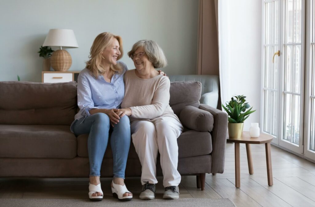 An older adult sits on a couch with their adult child. They embrace each other and smile.

