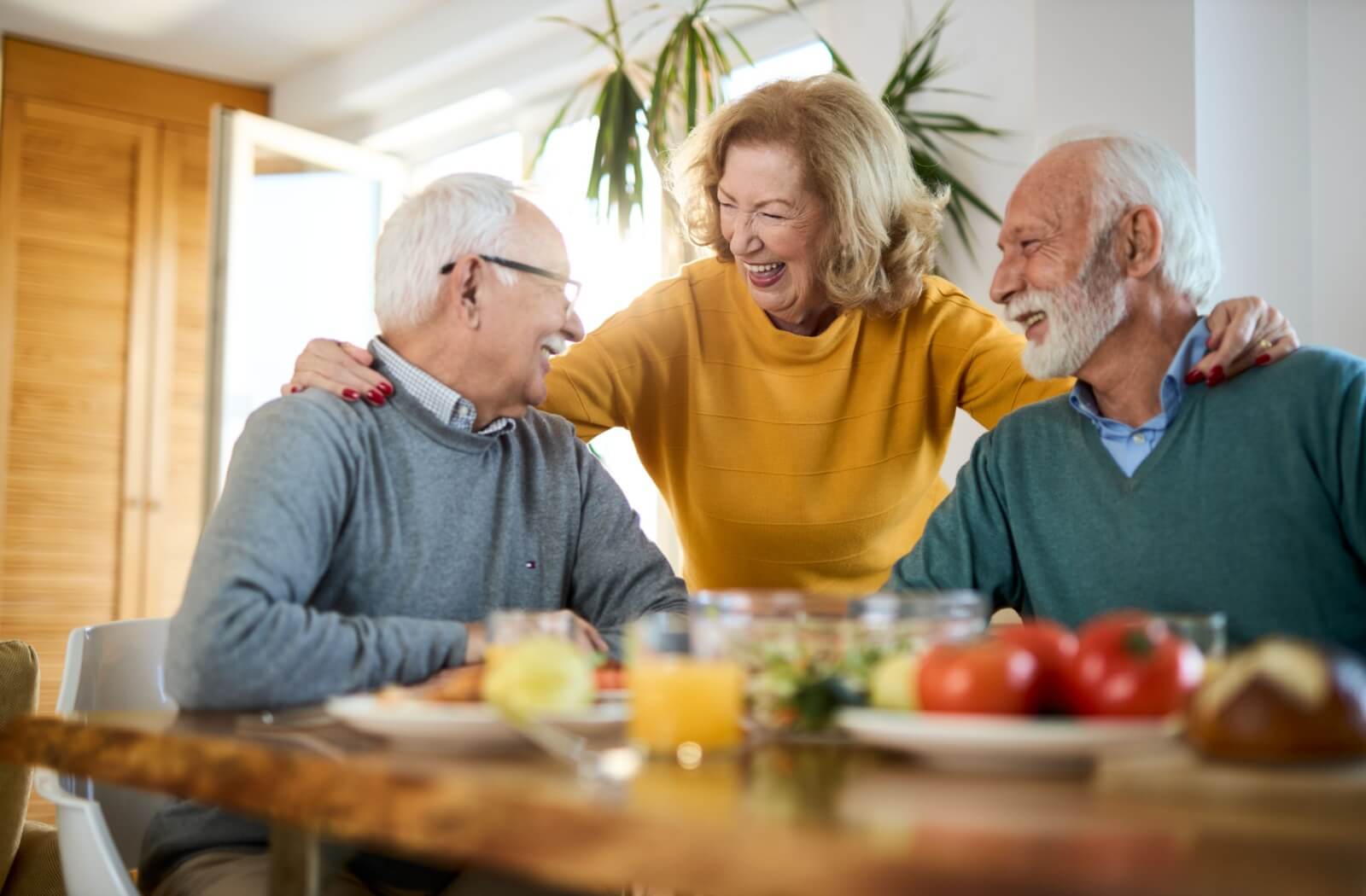 Three senior friends are having dinner together. One of the senior stands to put their arms around their two friends' shoulders and embrace them.