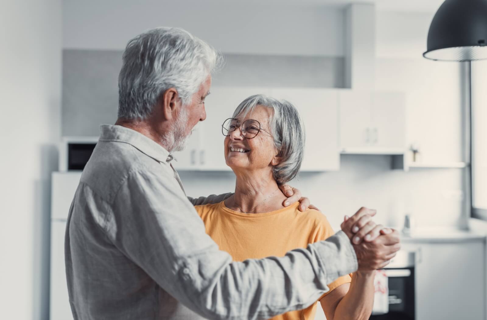 An older adult couple embrace each other closely and smile tenderly as they dance through their kitchen.