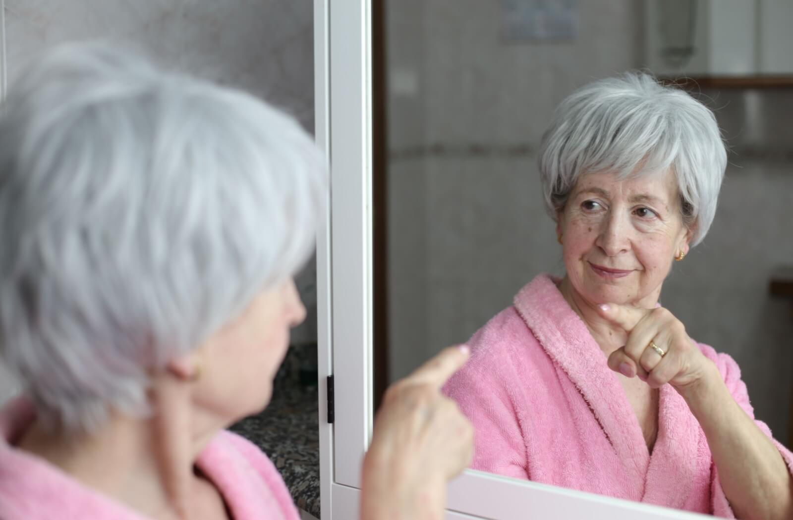 An older adult wearing a pink robe looks in the mirror and points at themselves, giving themselves a pep talk for the day.