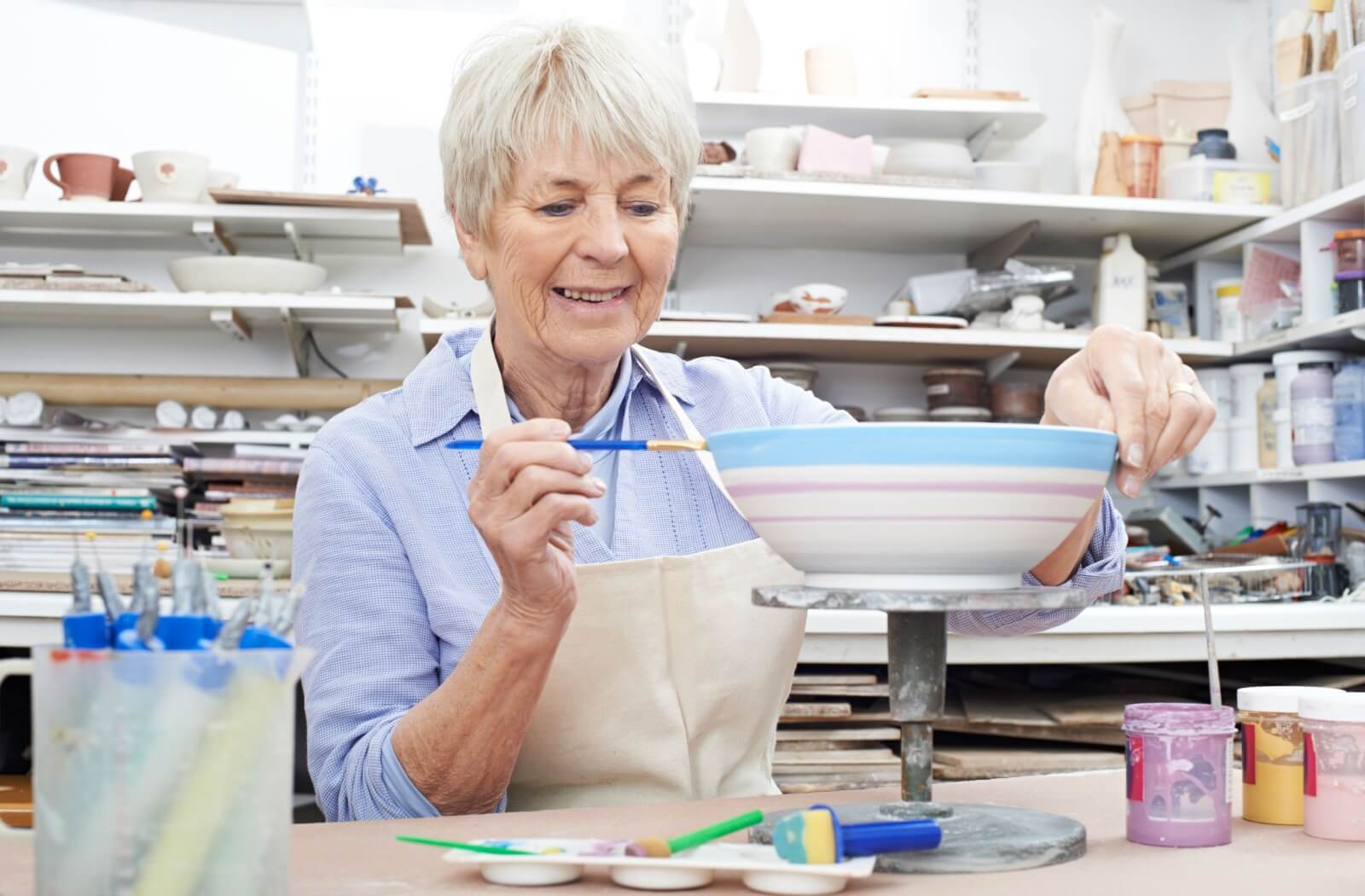 An older adult smiling while painting a handcrafted ceramic bowl in a home workshop.