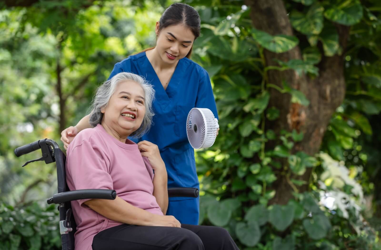 A caregiver holds a portable fan for an older adult while on a stroll outdoors in the summer.