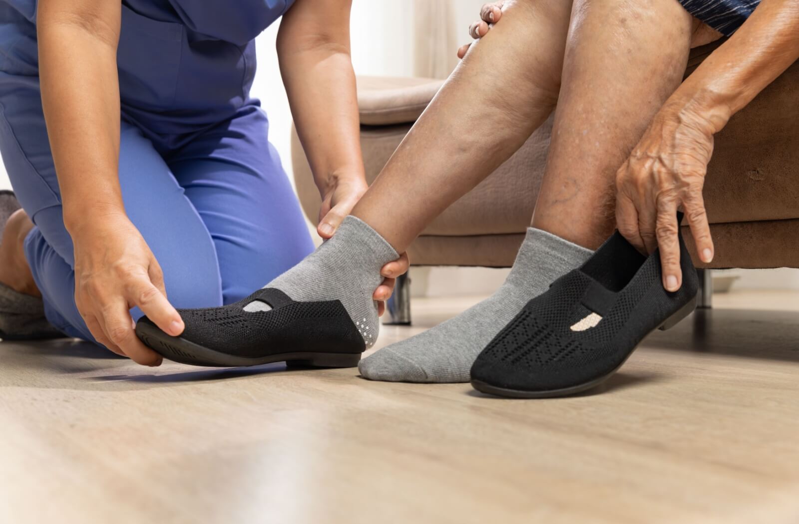 A kneeling caregiver helping an older adult put on a pair of comfortable black shoes.