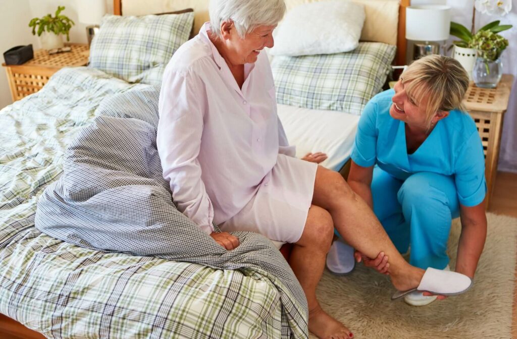An older adult with dementia smiling at a caregiver as they help them put on a comfortable pair of white slippers.