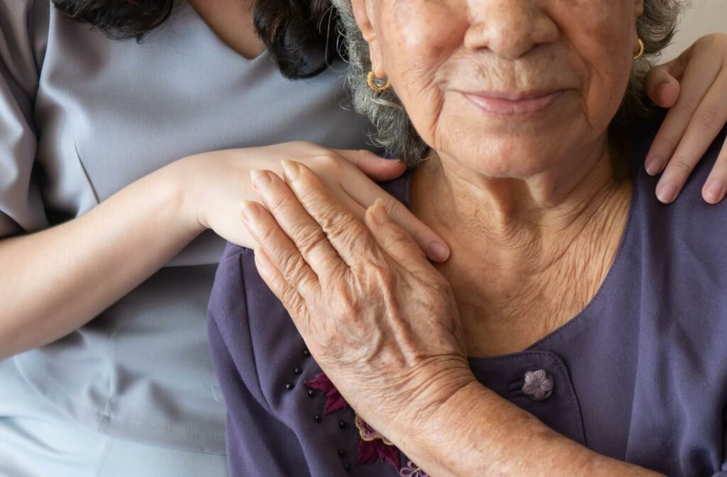 An older adult in a wheelchair smiling at the camera while a caregiver rests their hands on their shoulders.