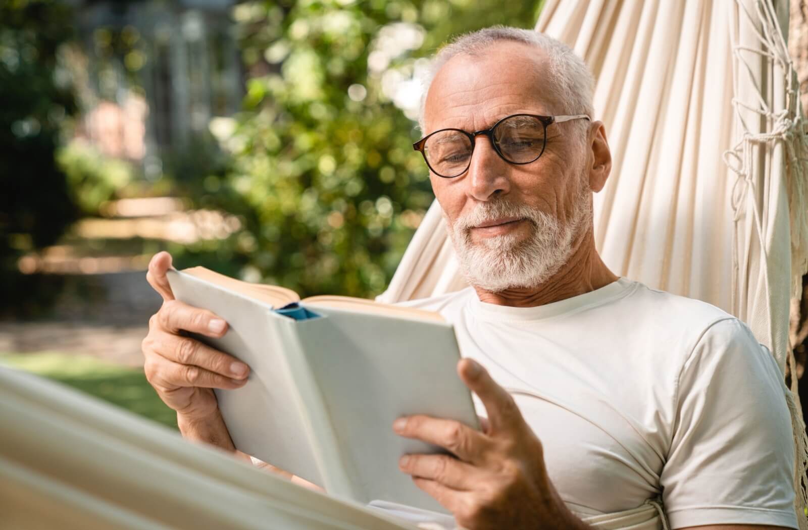 An older adult lying in a hammock on a summer day and reading a hardcover book.