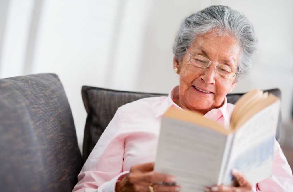 A smiling older adult sitting on their couch and reading a paperback novel.