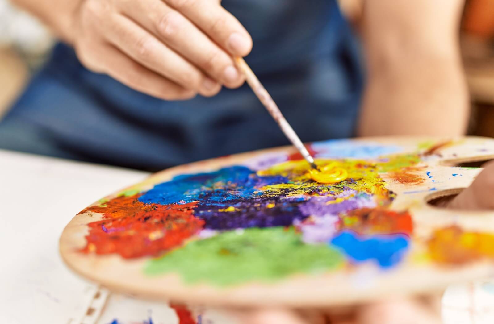 A close-up of an older adult dipping a paintbrush in paint on a palette covered in different colors.