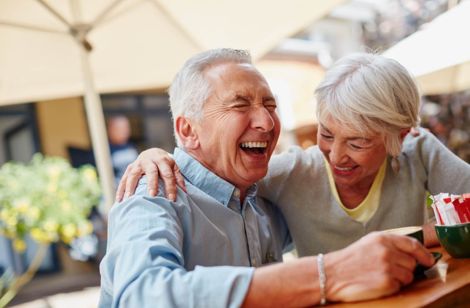An older couple laughing while sitting under an umbrella on a patio in senior living.