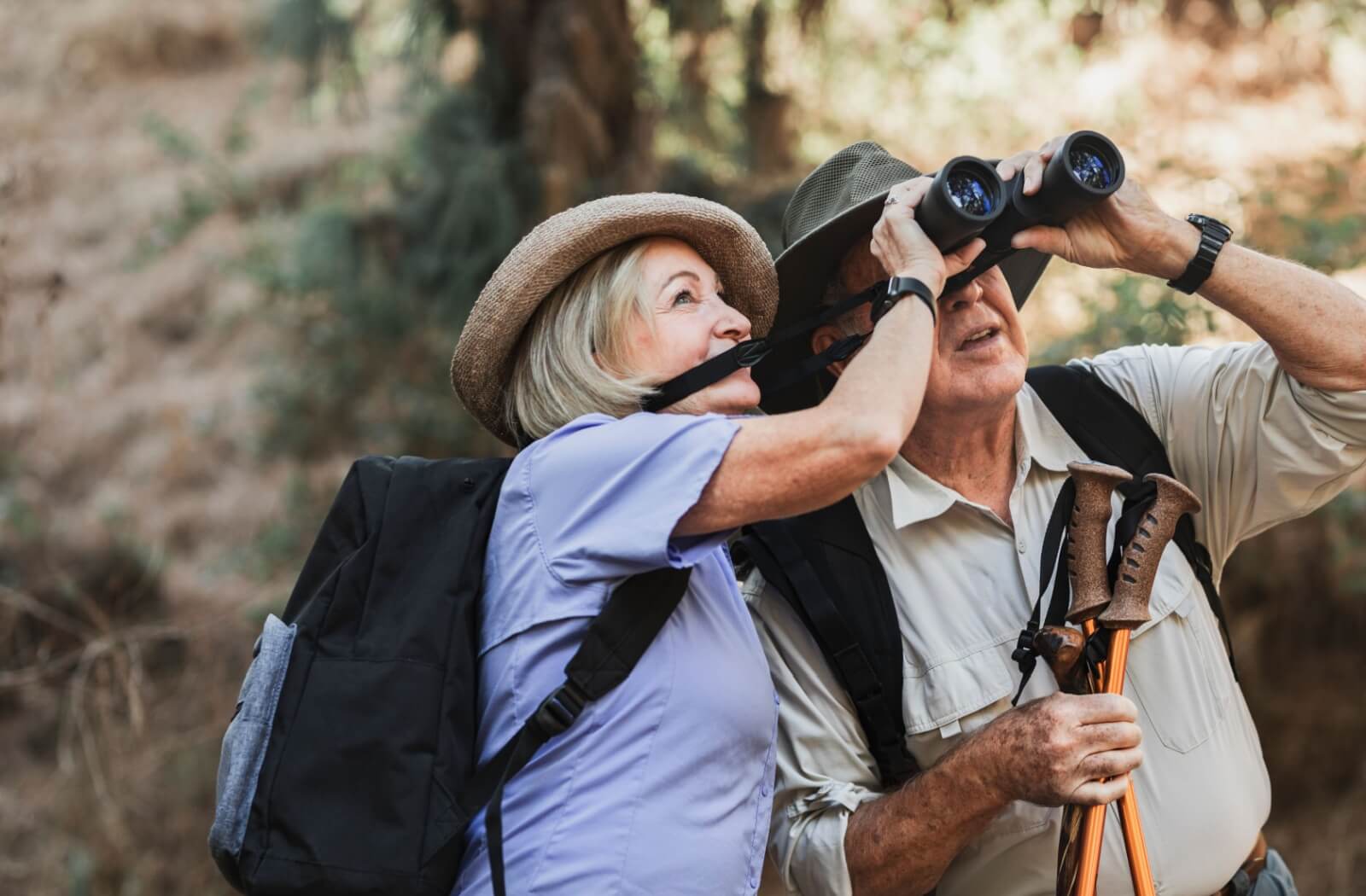 An older couple sharing a pair of binoculars while on a nature hike in the woods to birdwatch.