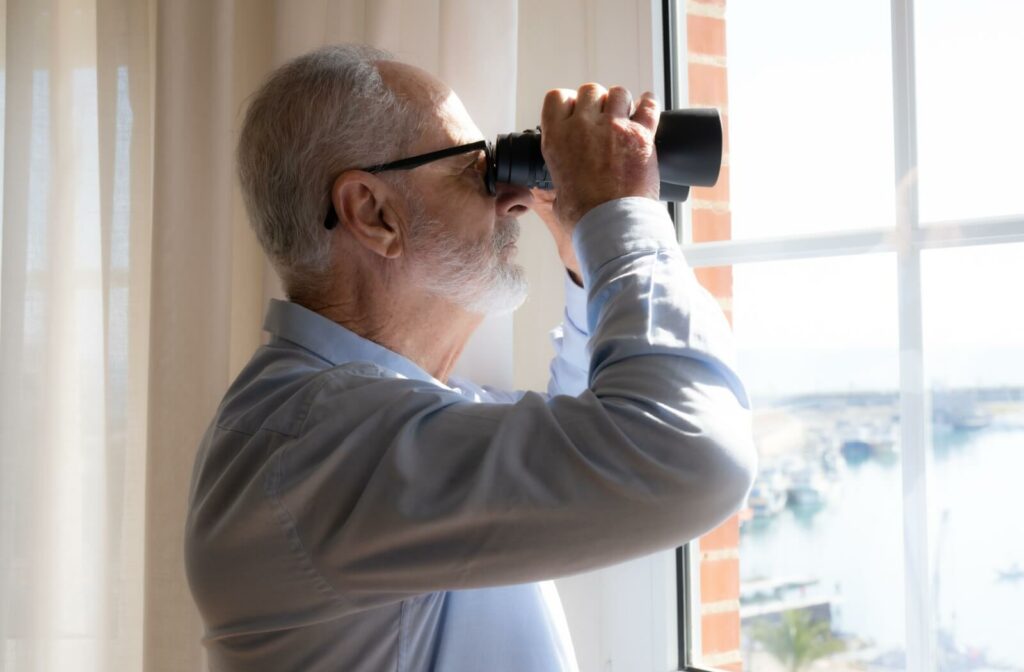 An older adult using a pair of binoculars to watch birds from their living room window.
