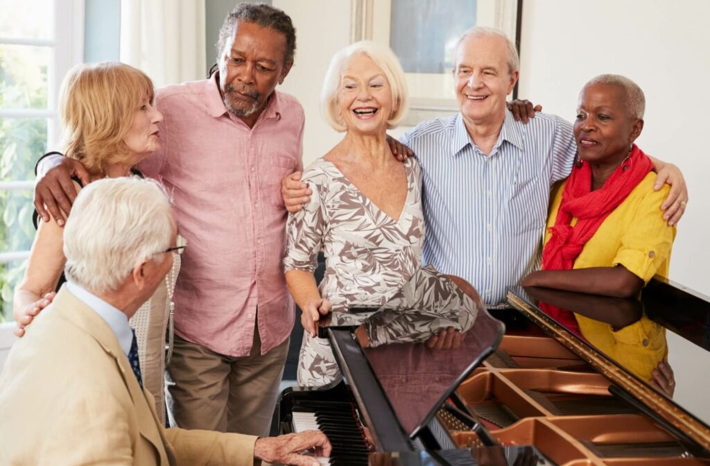 A group of older adults arm-in-arm around a piano in the common room of a senior living community, singing and smiling together.