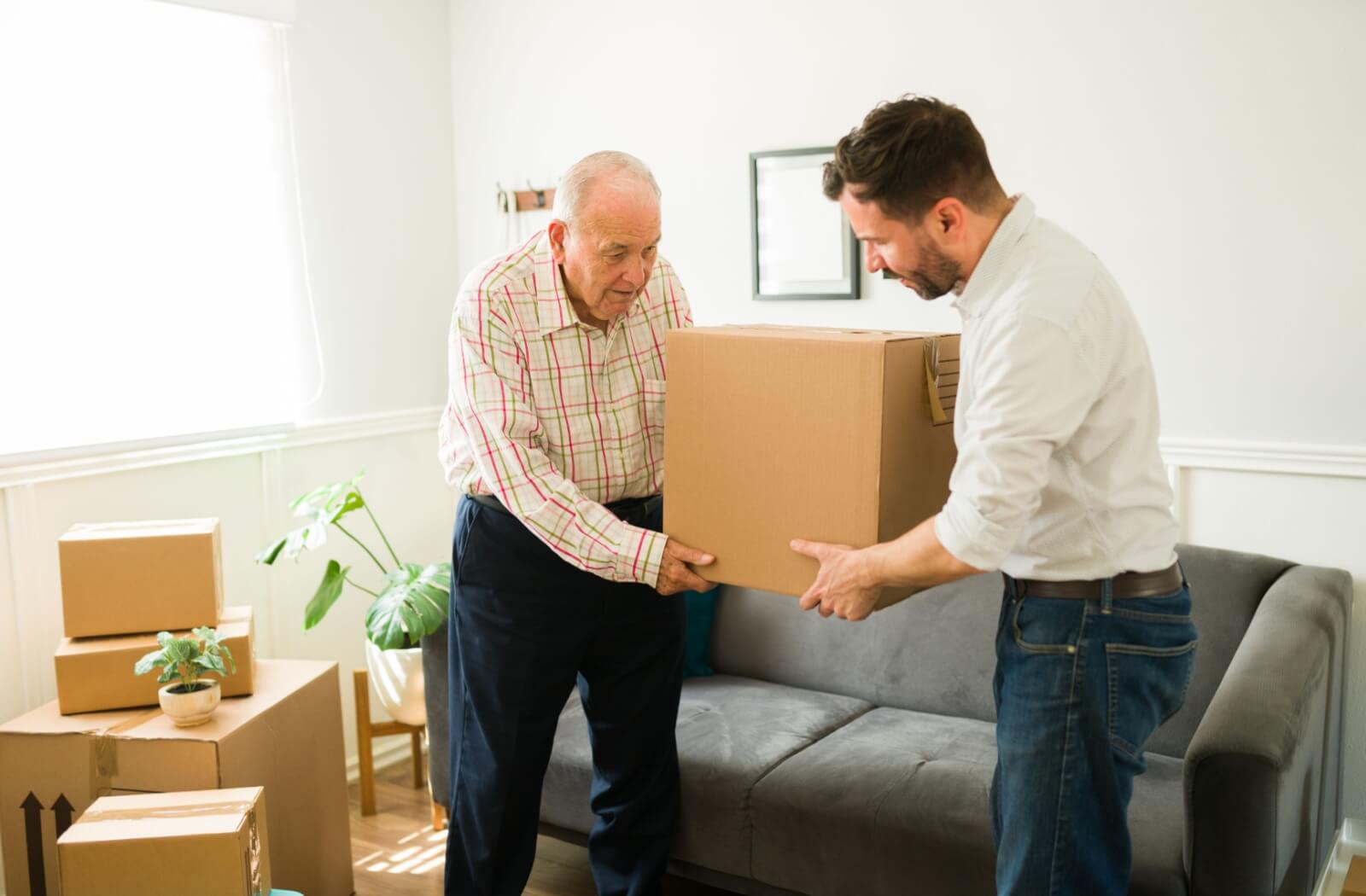 An adult child helping their older parent carry a box while downsizing their home for a move to assisted living.