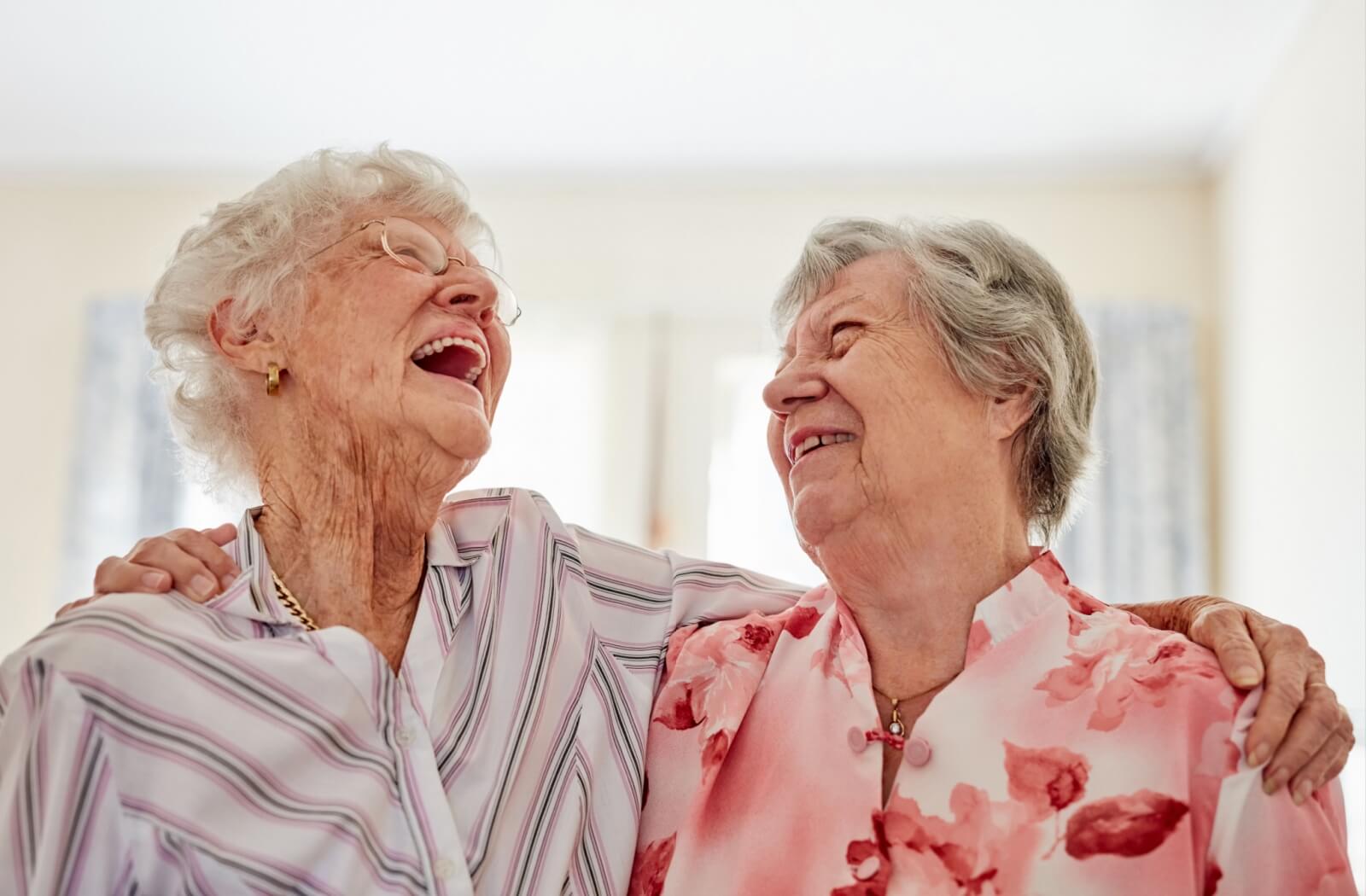 A pair of older adults in senior living standing with their arms around one another and laughing.