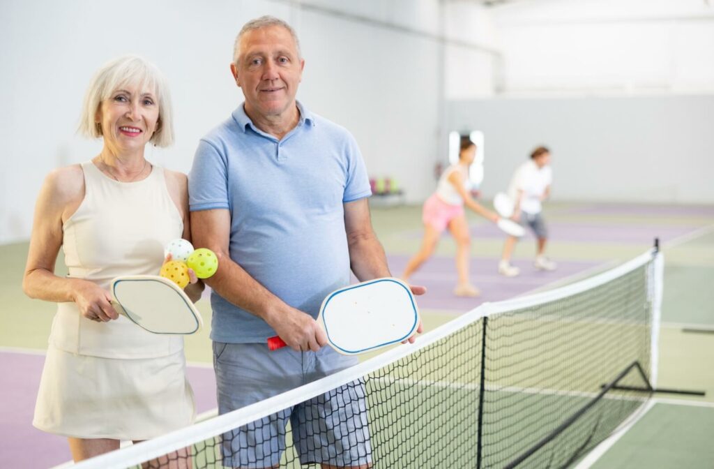 An older couple smiles while holding pickleball paddles in an indoor court
