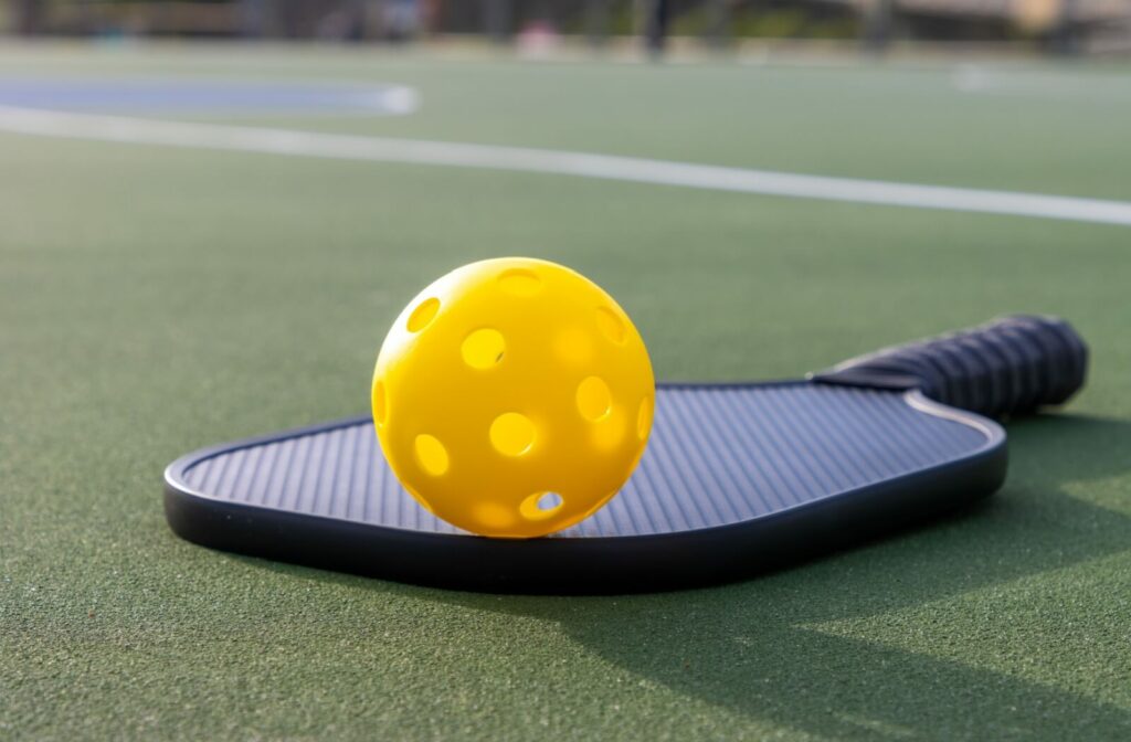 A close-up image of a pickleball sitting on a paddle lying on a green court