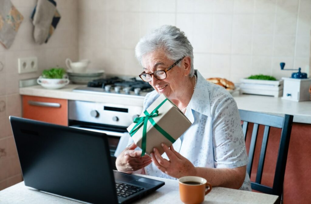 Older adult smiling while holding a wrapped gift during a video call on laptop at kitchen table with coffee