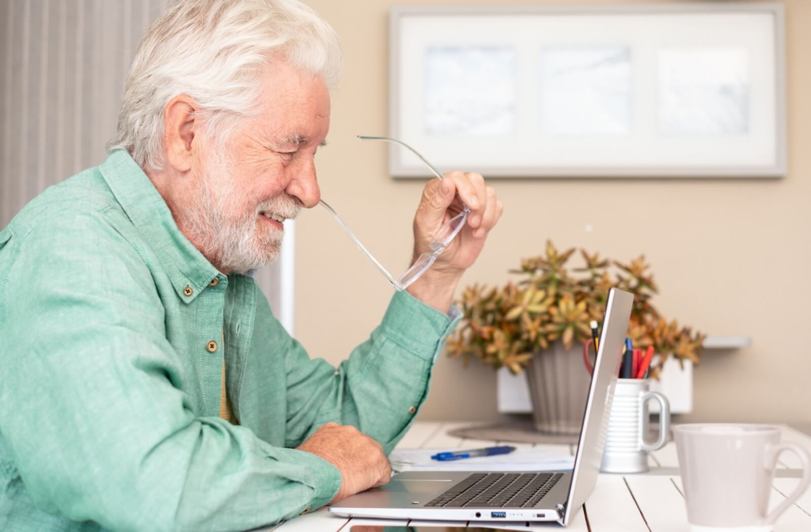 Smiling older adult in green shirt holding glasses while looking at laptop during an online remote learning class at home