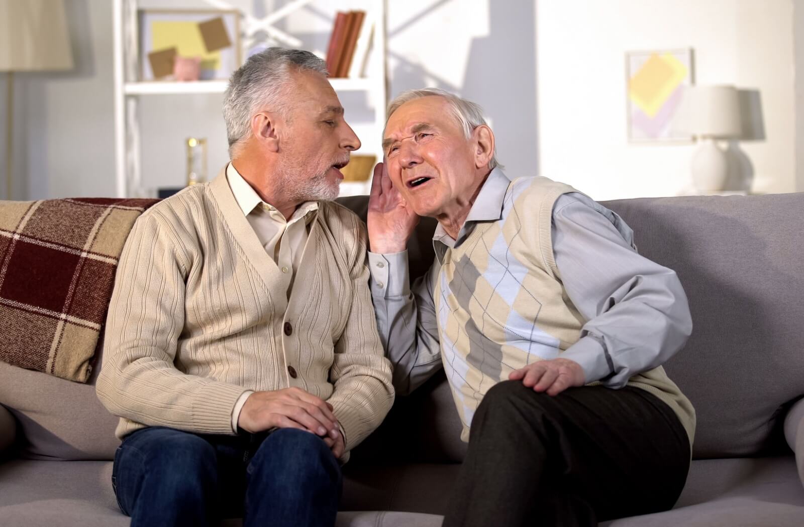 An older adult cups their hand around their ear and leans closer to their friend on a couch to hear, indicating hearing loss