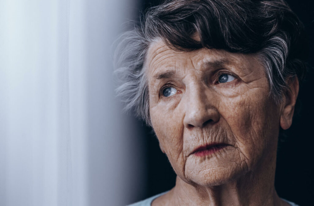 An elderly woman stands by the window, gazing outside with a lonely expression on her face.