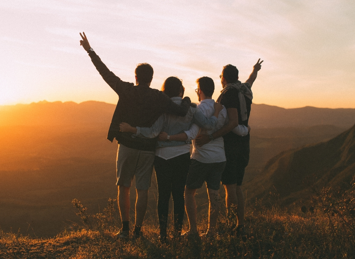 A group of friends standing in front of a sunset, with their hands up and experiencing happiness