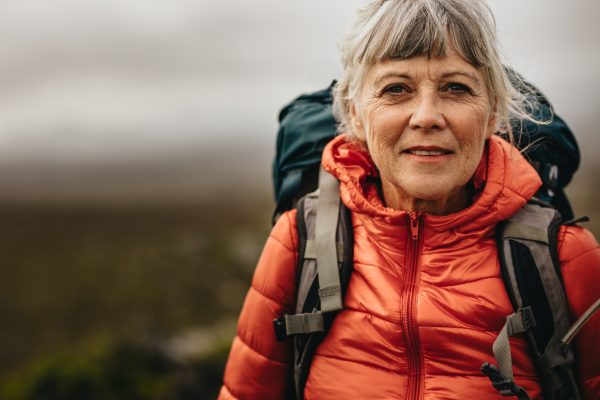 Portrait of a senior woman standing outdoors on a holiday. Senior woman wearing a backpack and jacket standing during a trekking trip.